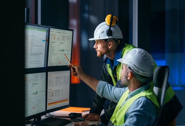 Two factory workers wearing helmets, looking at screens of data